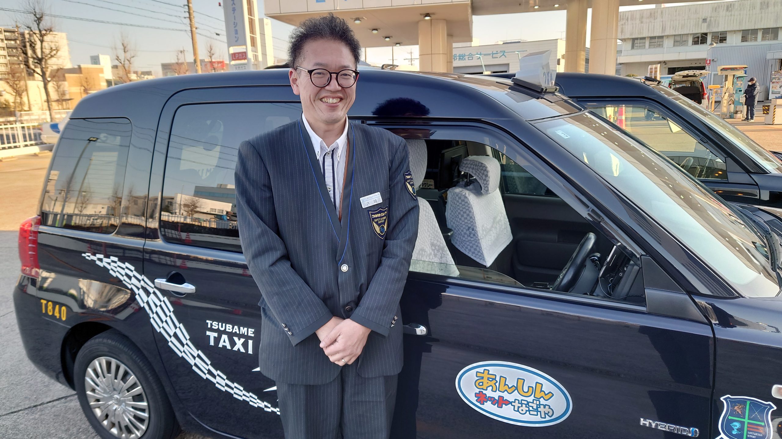 名古屋市で働くつばめタクシーの日本人ドライバーが笑顔で車の前に立つ様子。安定した給与と働きやすさを象徴するシーン。